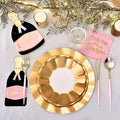 The Sophistiplate Kiss Me at Midnight Table Set includes designer disposable plates, pink and gold cutlery, "Kiss me at midnight" napkins, champagne bottle decorations, candles, and gold foliage on a white tablecloth.