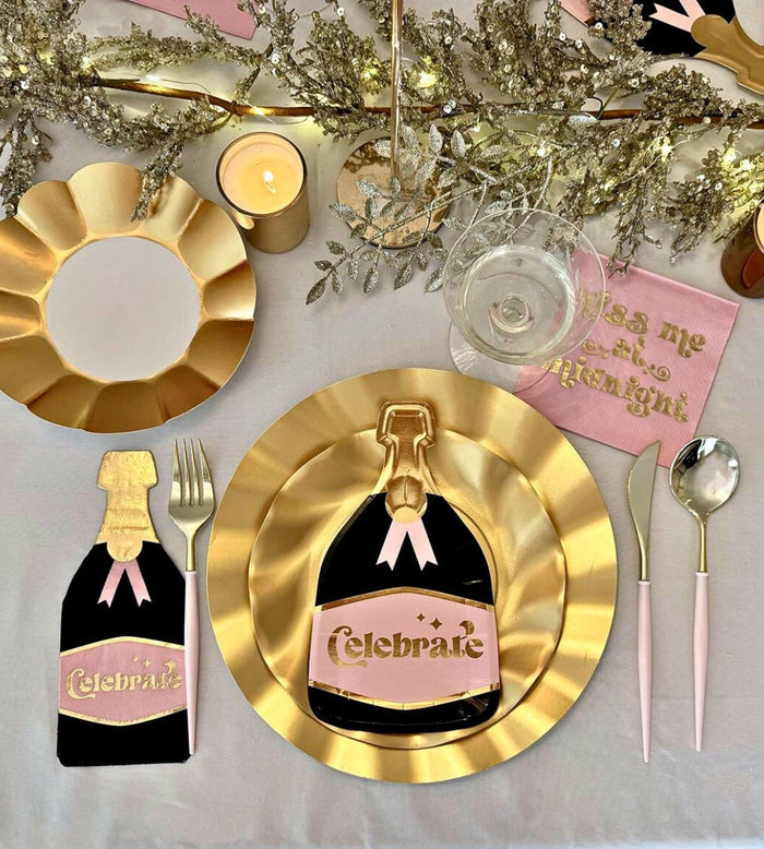 The Kiss Me at Midnight Table Set by Sophistiplate includes designer gold-rimmed plates, “Celebrate” cookies, gold and pink utensils, a candle, champagne glass, and sparkling gold accents atop a crisp white tablecloth.