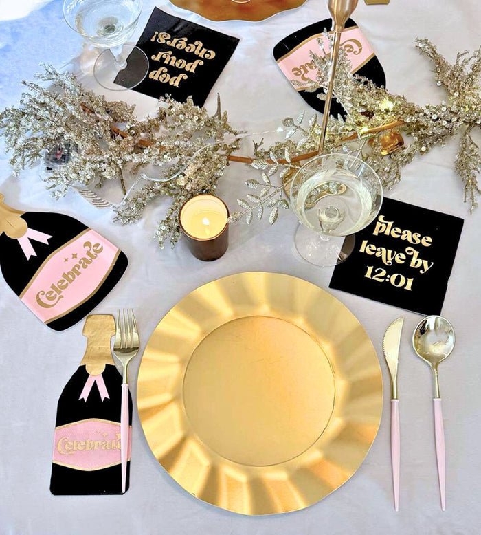 The Sophistiplate Kiss Me at Midnight Table Set includes a gold plate, gold utensils, champagne bottle napkins, "Celebrate" and "please leave by 12:01" black and gold coasters, white wine glasses, and sparkly silver decorations for a festive look.