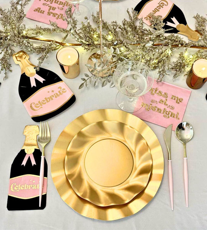 The Sophistiplate Kiss Me at Midnight Table Set includes designer disposable plates, pink cutlery, Celebrate bottle-shaped napkins, Kiss me at midnight pink napkins, gold foliage, candles, and fairy lights for a festive tablescape.