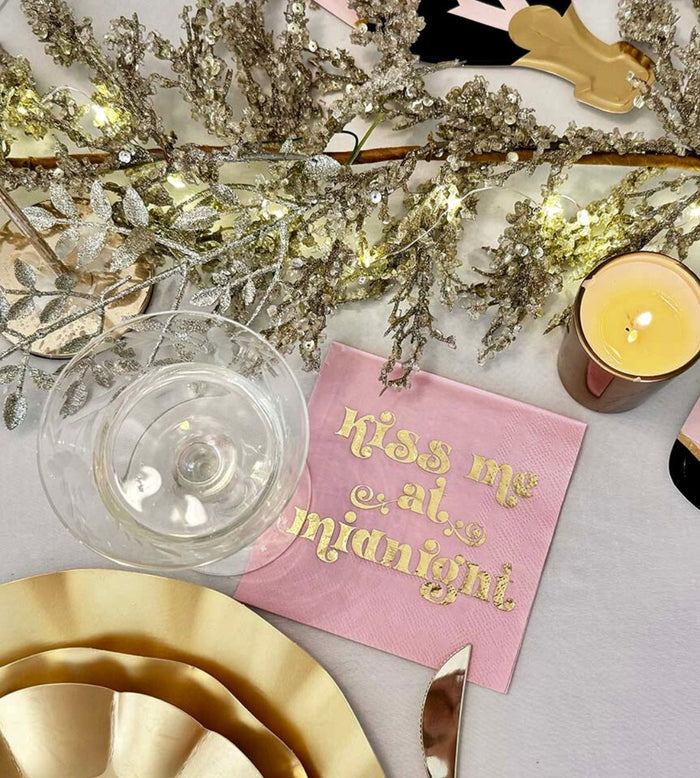 The Sophistiplate Kiss Me at Midnight Table Set features designer disposable plates, a wine glass, lit candle, decorative silver lighted branches, and a pink napkin with "Kiss me at midnight" in gold. Perfect for festive tablescapes-in-a-box.