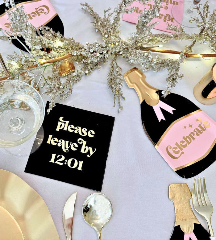 The Kiss Me at Midnight Table Set by Sophistiplate includes gold and pink designer disposable plates, Celebrate decor, a champagne bottle cutout, silverware, a glass, and a black "please leave by 12:01" napkin.