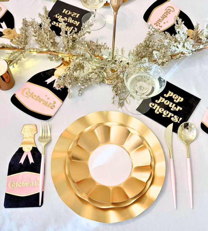 The Kiss Me at Midnight Table Set by Sophistiplate includes designer disposable plates, pink-handled cutlery, champagne flutes, black napkins with “pop pour cheers!” in gold, and bubbly bottle décor on a white tablecloth with gold foliage.