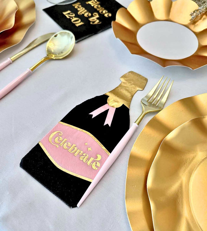 The Kiss Me at Midnight Table Set by Sophistiplate includes gold designer disposable plates, cutlery, pink-handled utensils, a champagne bottle “Celebrate” napkin, and a crisp white tablecloth for a festive tablescape.