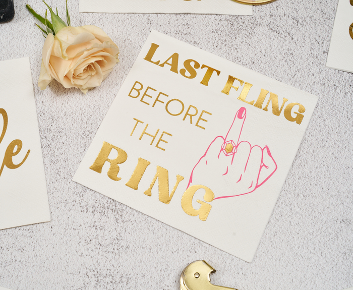Sugarplum's Last Fling before the Ring Cocktail Napkins (26pk) feature gold script and a pink hand with an engagement ring, perfect for bachelorette parties or bridal showers, shown next to a pale rose on a light background.