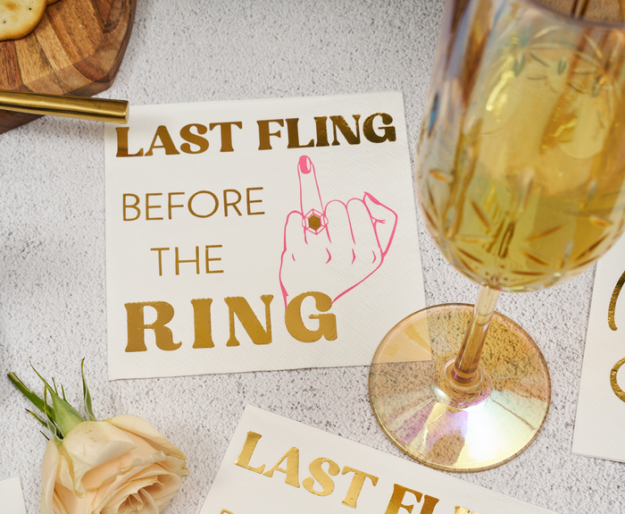 Sugarplum’s Last Fling before the Ring Cocktail Napkins (26pk) feature gold text and a pink hand flipping the bird—plus fun accents like a champagne glass, peach rose, and snacks—perfect for your unforgettable bachelorette party!.