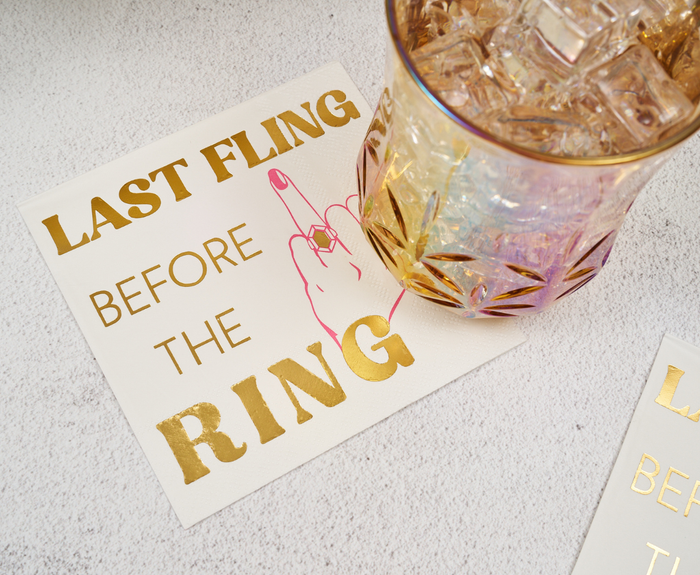 A glass of ice rests on Sugarplum's "Last Fling before the Ring" cocktail napkins (26pk), featuring gold accents and a pink hand with a ring illustration.