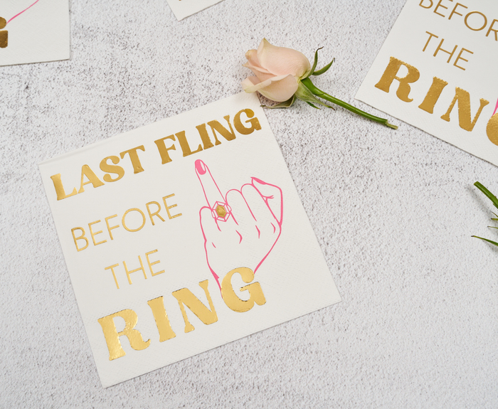 Sugarplum Last Fling before the Ring Cocktail Napkins (26pk) feature "LAST FLING BEFORE THE RING" and a pink hand with a ring—perfect bachelorette party napkins displayed beside a light pink rose on a textured white surface.