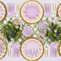 The Lavender Fields Table Setting by Sophistiplate LLC features designer lavender plates, gold utensils, pink napkins, and lush green-white floral centerpieces on a white tablecloth with a green lattice runner.