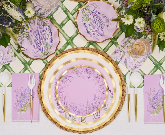 The Lavender Fields Table Setting by Sophistiplate LLC features designer disposable plates in lavender, gold cutlery, pink floral napkins, woven placemats, and floral centerpieces on a green and white lattice tablecloth.
