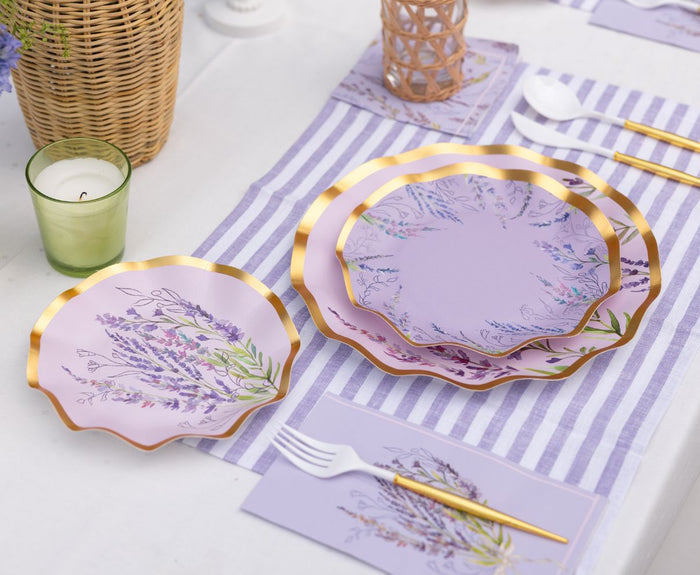A close-up of the Lavender Fields Table Setting by Sophistiplate LLC, featuring scalloped lavender plates with gold rims, floral accents, striped placemats, gold cutlery, a green candle, and a wicker container on white linen.