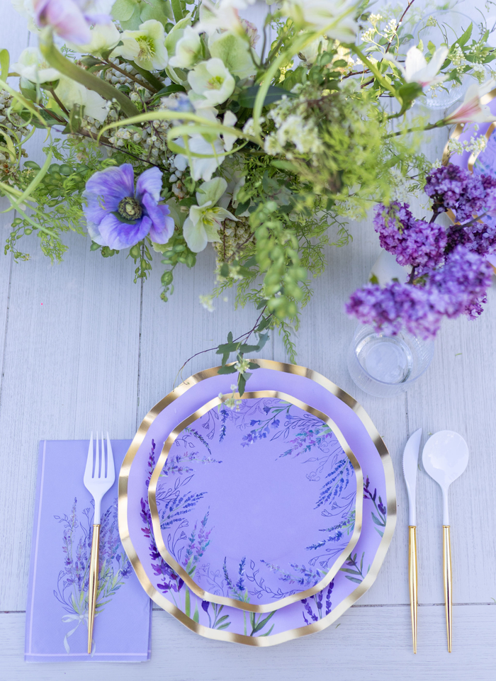The Lavender Fields Table Setting by Sophistiplate LLC features designer disposable plates, gold-rimmed utensils, and napkins, accented by a floral centerpiece of purple and white blooms on a white wooden table.