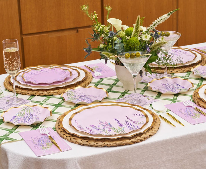 The Lavender Fields Table Setting by Sophistiplate LLC seats four with designer pink floral plates, purple napkins, gold cutlery, woven placemats, and glassware. A white and green floral centerpiece completes the look.
