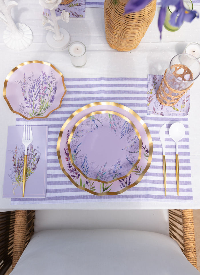 The Lavender Fields Table Setting by Sophistiplate LLC features designer disposable plates with lilac florals and gold edges, purple striped placemat, gold cutlery, floral napkins, wicker glass holder, candles, and flower basket—perfect for your party-in-a-box.