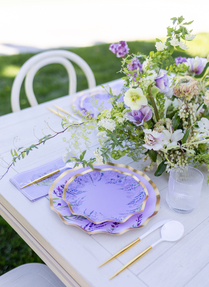The Lavender Fields Table Setting by Sophistiplate LLC features gold-rimmed designer plates, gold cutlery, a clear glass, white spoon, and a floral centerpiece with purple, white, and green accents for an elegant outdoor table.