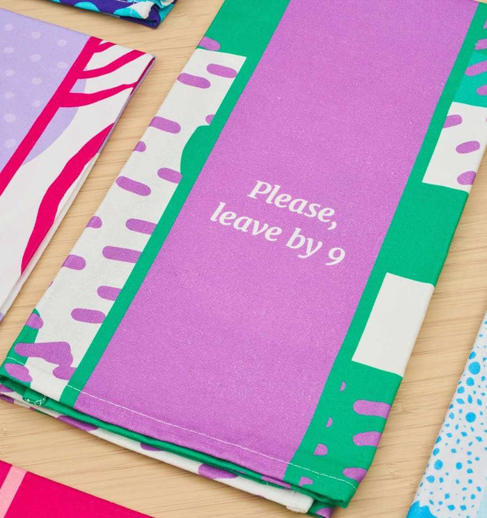 The Leave by 9 Dish Towel from Drinks On Me features a bold, colorful abstract print in purple, green, and white with "Please, leave by 9" at the center—great for gifting or pairing with a sarcastic napkin. Shown on a light wooden surface.