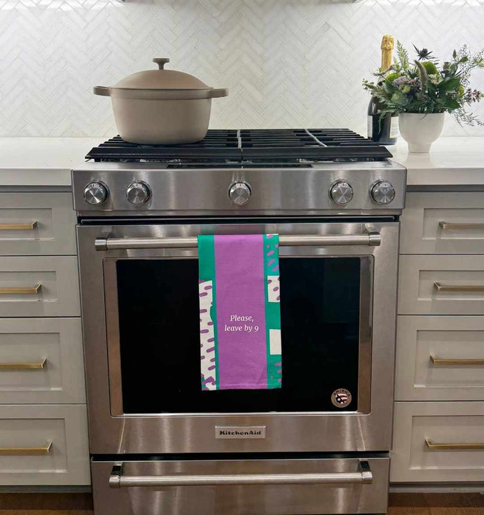 The Leave by 9 Dish Towel by Drinks On Me hangs on a modern stainless steel oven, featuring the funny message “Please, leave by 9”—a perfect humorous kitchen gift. A tan pot, flower arrangement, and bottle complete the stylish scene.