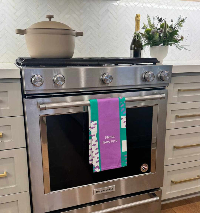 A modern kitchen showcases the Drinks On Me Leave by 9 Dish Towel on a stainless steel oven door. A pot, bottle, and flower arrangement rest on the white countertop above.