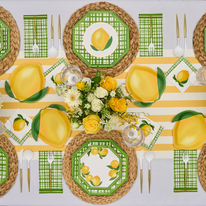 The Lemon and Stem Table Setting by Sophistiplate serves four with designer lemon-themed disposable plates, green plaid napkins, woven placemats, white and gold cutlery, and a centerpiece of yellow roses and white flowers on a yellow-striped runner.