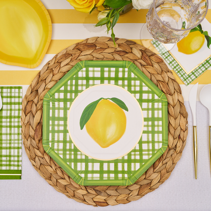 The Lemon and Stem Table Setting by Sophistiplate includes lemon-illustrated designer plates, woven placemat, lemon napkin, clear glass, white cutlery, and a yellow-and-white striped tablecloth with yellow flowers.