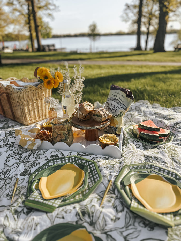 A picnic on a patterned blanket by the lake features Sophistiplate’s Dinner Plate Lemon and Stem/8pk, summer tableware, sunflowers, chips, watermelon slices, drinks, food, and a wicker basket in a scenic park setting.