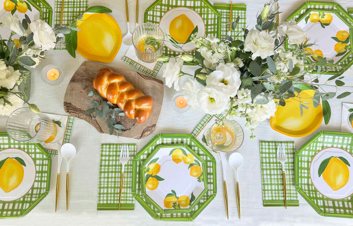 A vibrant table features green plaid placemats, Sophistiplate Salad Plate Lemon and Stem/8pk, gold utensils, white napkins, vases of white flowers, candles, and a braided loaf on a wooden board—perfect for summer gatherings.