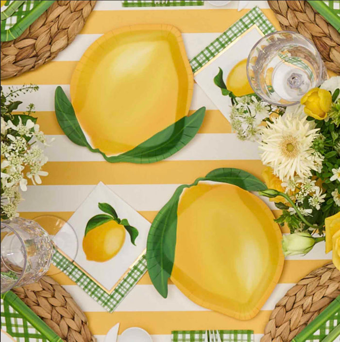 The Sophistiplate Lemon and Stem Table Setting includes designer lemon-themed plates, a yellow and white striped tablecloth, green plaid napkins, woven placemats, clear glasses, and coordinating white and yellow floral arrangements.