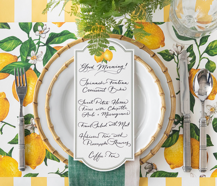 A breakfast table is set with Hester & Cook's lemon-themed placemats, bamboo-edged plates, and Lemon Table Card - Pack of 12, plus a handwritten menu featuring spinach frittata, croissant bake, fruit salad, hibiscus tea, and coffee.
