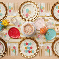 The Let’s Celebrate Table Setting by Sophistiplate includes designer disposable plates, colorful balloon motifs, confetti cake decor, bright flowers, party napkins with “HOORAY,” and a gold and white striped table runner for a festive tablescape.