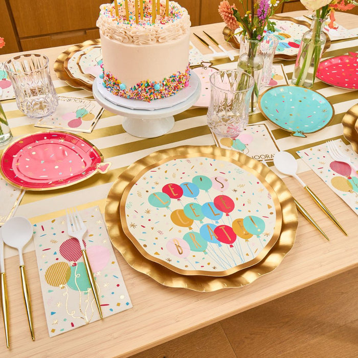 The Sophistiplate Let's Celebrate Table Setting includes designer disposable plates, balloon napkins, gold cutlery, glasses, candles and sprinkles for cake, and flower vases—perfect for a festive birthday tablescape on a light wood table.