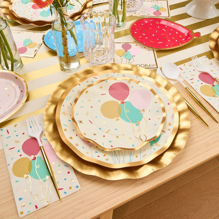 The Let's Celebrate Table Setting by Sophistiplate includes balloon-print plates, gold-rimmed chargers, a striped tablecloth, festive napkins, clear glasses, white cutlery, and flower-filled mini vases for a colorful party tablescape.