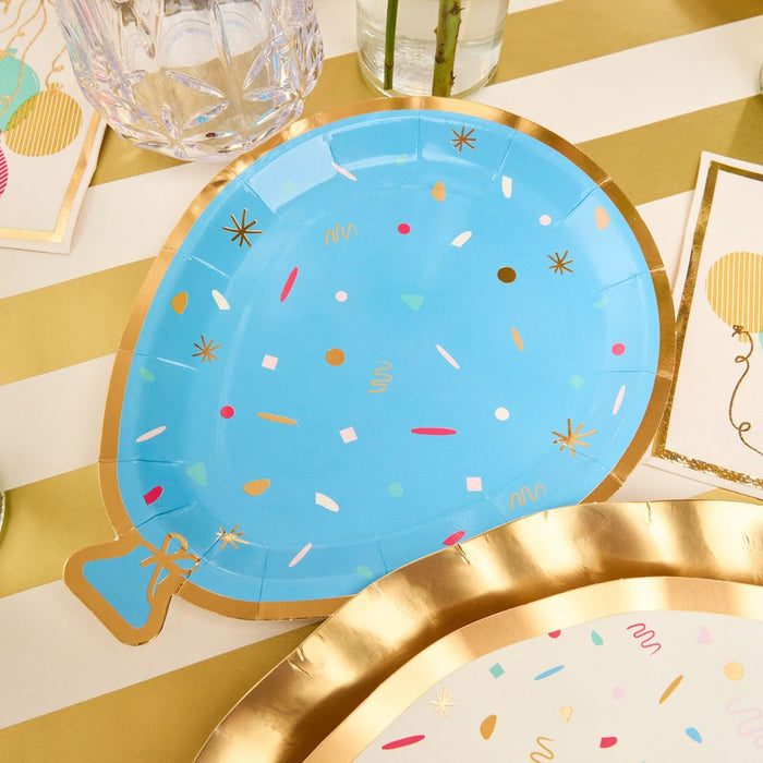 The Sophistiplate Let's Celebrate Table Setting features blue balloon-shaped plates with confetti designs, paired with balloon napkins, a gold-rimmed party plate, and a drink on a gold and white striped table.