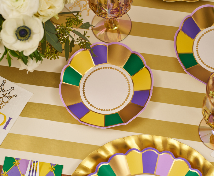 Celebrate with the Let's Mardi Table Setting by Sophistiplate: designer paper plates in purple, green, gold, and yellow atop a gold-striped tablecloth plus matching party plates, gold cutlery, and festive details for a vibrant Mardi Gras tablescape.