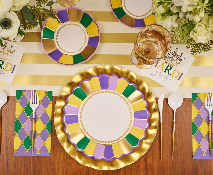 The Let's Mardi Table Setting from Sophistiplate features designer paper plates in green, purple, and gold with crown napkins, gold utensils, a striped runner, wine glasses, and white flower centerpieces for a festive disposable tablescape.