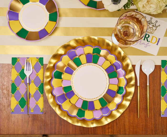 The Let's Mardi Table Setting by Sophistiplate features designer plates in gold, green, and purple, a striped placemat, vibrant napkins, white cutlery, a drink cup, and a “Mardi Gras” card with a crown—perfect for your festive celebration.