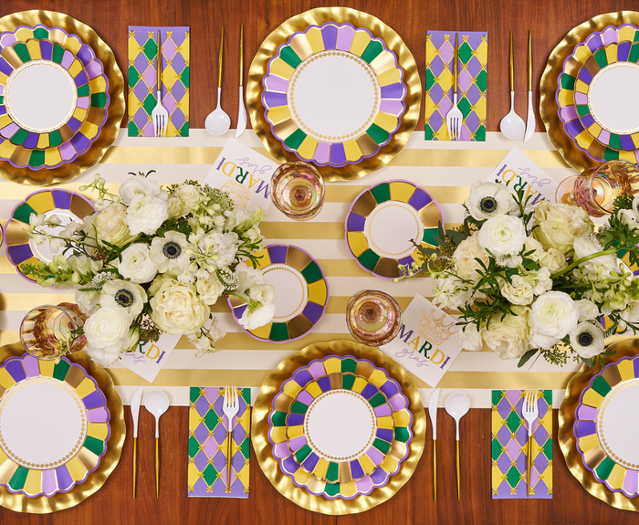 A festive Mardi Gras table set with gold, purple, and green plates, gold chargers, white flowers in vases, Sophistiplate Let's Mardi Cocktail Napkins (26pk), gold utensils, “Mardi” cards, and a striped table runner.