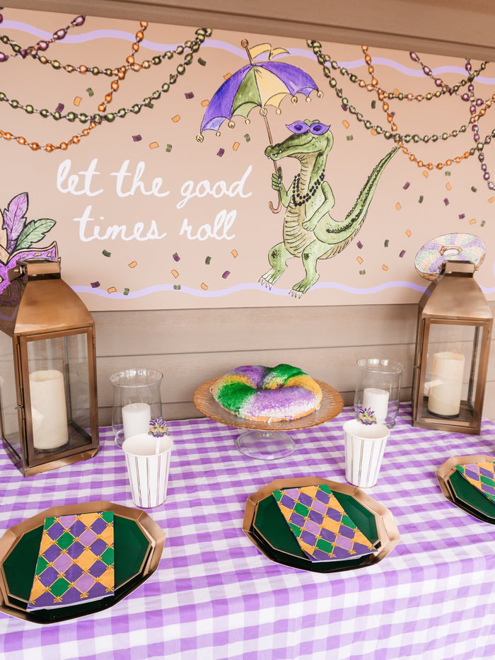 A Mardi Gras table featuring Sophistiplate’s “Let's Mardi” guest towels, purple, green, and gold plates, king cake, candles in lanterns, and a mural of an alligator holding an umbrella with the phrase “let the good times roll.”.