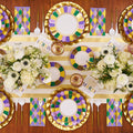 The Let's Mardi Table Setting by Sophistiplate features designer paper plates in gold, purple, and green, gold utensils, floral centerpieces, argyle napkins, “Mardi Gras” cards, and a gold-striped runner for a stunning disposable tablescape.