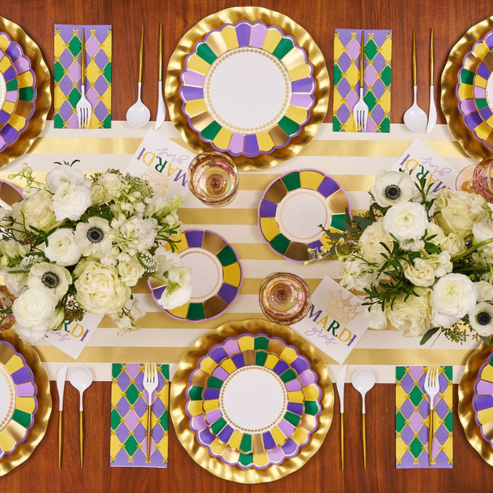 The Let's Mardi Table Setting by Sophistiplate features designer paper plates in gold, purple, and green, gold utensils, floral centerpieces, argyle napkins, “Mardi Gras” cards, and a gold-striped runner for a stunning disposable tablescape.