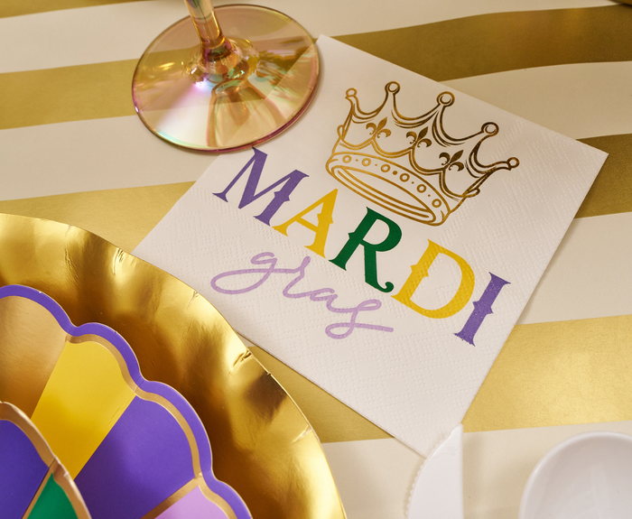 The Let's Mardi Table Setting by Sophistiplate features festive napkins with gold crowns, designer plates, and an iridescent glass on a striped gold and white tablecloth for a vibrant disposable Mardi Gras tablescape.