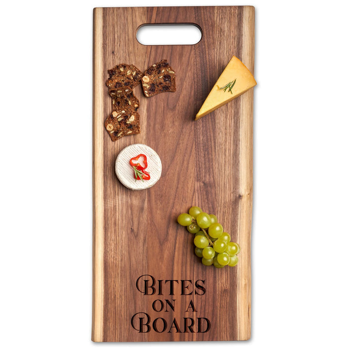 The Maple Live Edge Walnut Single Handle Charcuterie Board | Parisian Bites On Board (24" x 12") displays a cheese wedge, a small round cheese with pepper slice, crisps, and green grapes.