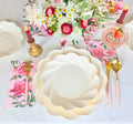The Love in Bloom Table Setting by Sophistiplate includes scalloped designer plates, gold and pink cutlery, floral napkins, red candles, a crystal glass with a strawberry, and a white and pink bouquet on a white tablecloth.