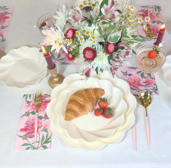 The Love in Bloom Table Setting by Sophistiplate features a croissant and strawberries on designer plates, with floral napkins, gold and pink cutlery, candles, a dessert glass, and a pink-white flower bouquet on a white tablecloth.