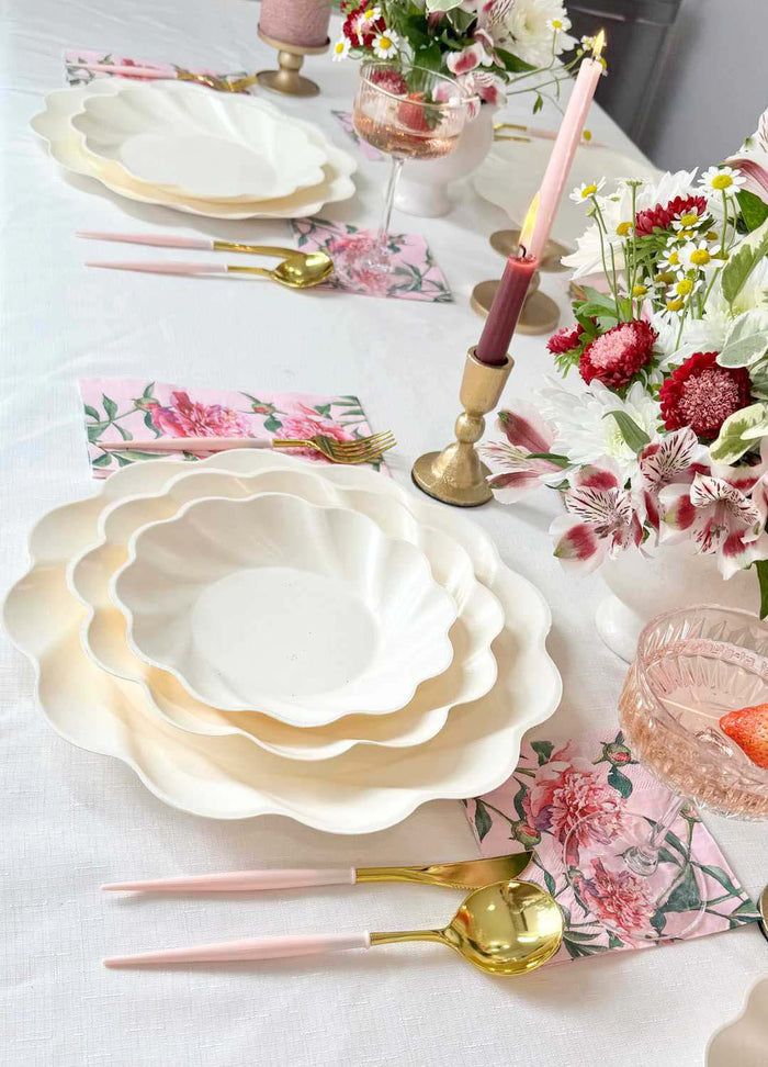 The Sophistiplate Love in Bloom Table Setting features a white tablecloth, floral-themed tablescape-in-a-box, scalloped designer plates, gold and pink cutlery, floral napkins, candles with holders, and vases of red, white & pink flowers.