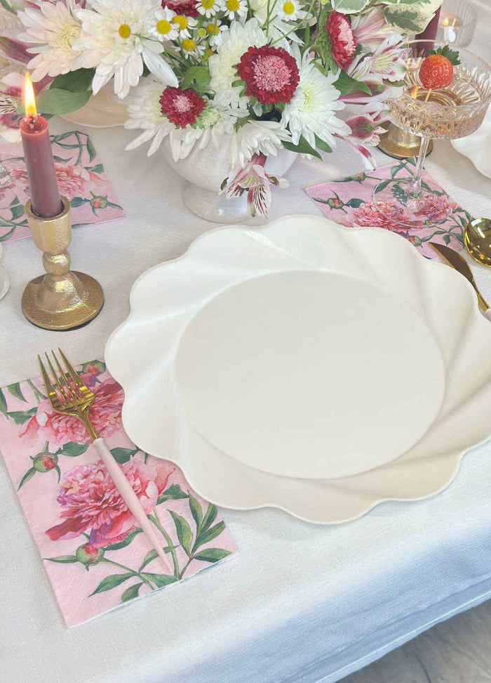 The Love in Bloom Table Setting by Sophistiplate features a white scalloped designer disposable plate on a floral napkin, gold fork, pink candle in a gold holder, champagne glass, and white-pink flower bouquet on a white tablecloth.