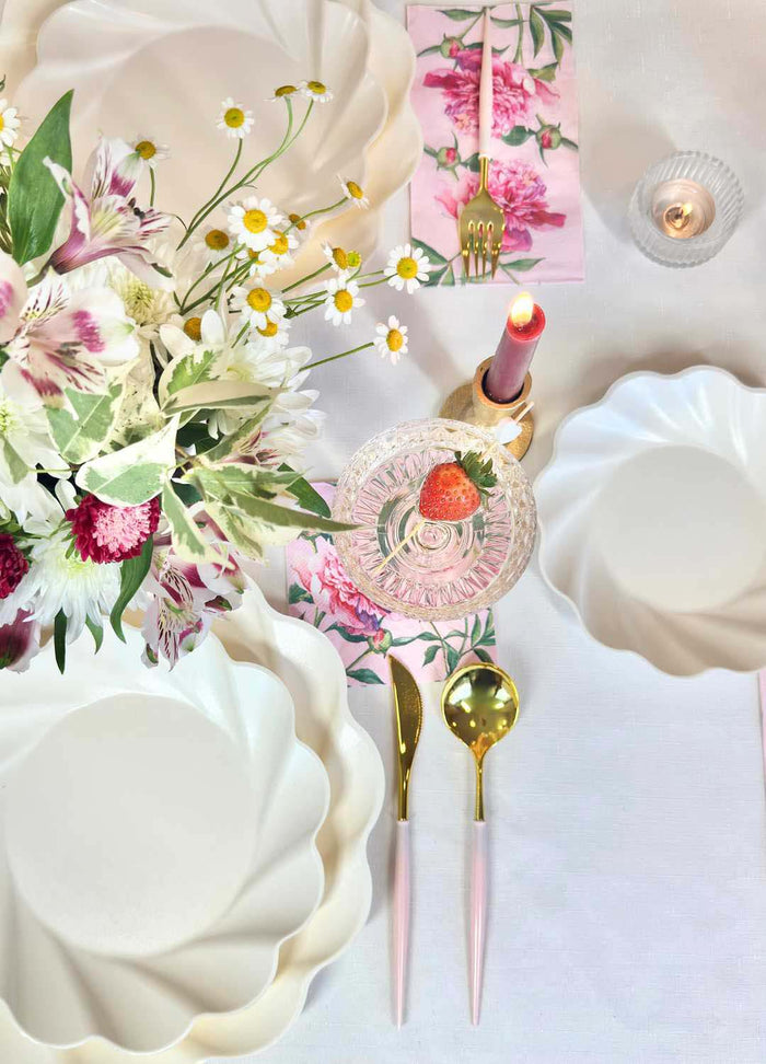 The Love in Bloom Table Setting by Sophistiplate features designer disposable plates, gold and pink cutlery, a floral napkin, pink candle, glass dessert dish with strawberry, bouquet of flowers, and lit tealight for an elegant spring tablescape.