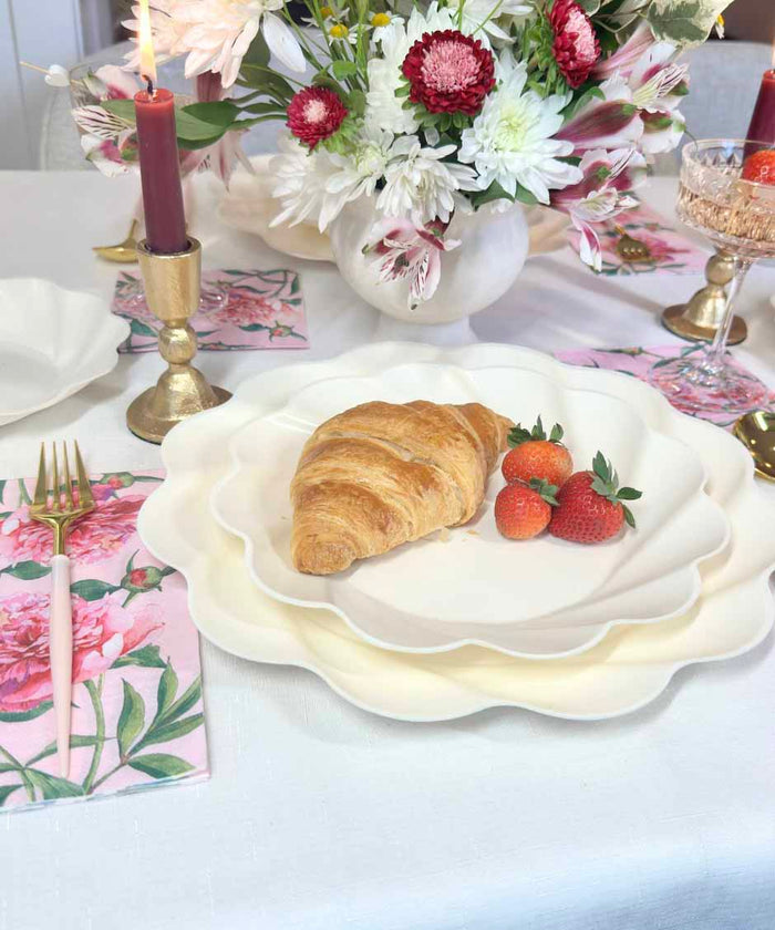 The Love in Bloom Table Setting by Sophistiplate features a scalloped designer plate with a croissant and strawberries, surrounded by floral napkins, gold cutlery, red candles, and a flower-filled vase for an elegant tablescape-in-a-box look.
