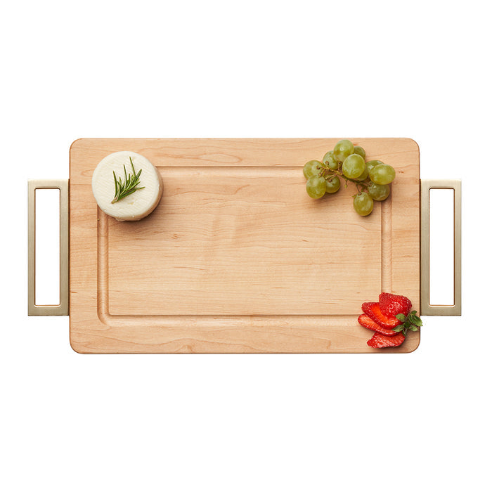 The Maple Wood Cutting + Cheeseboard (13 x 8", gold handles) by Maple is arranged with a round cheese wheel garnished with herbs, green grapes, and sliced strawberries for a beautiful presentation.