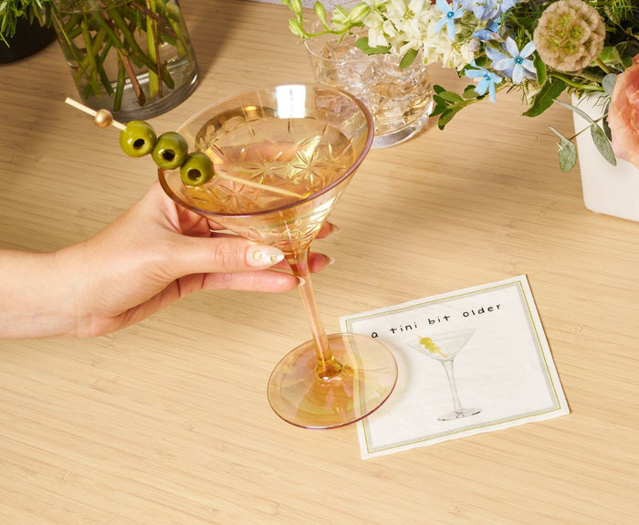 A hand holds a shatterproof Sophistiplate LLC Martini Classic Gold 8 Oz glass with three olives, next to an "a tini bit older" napkin. Flowers and a glass of ice sit on the wooden table. Sold as a 4 pack.