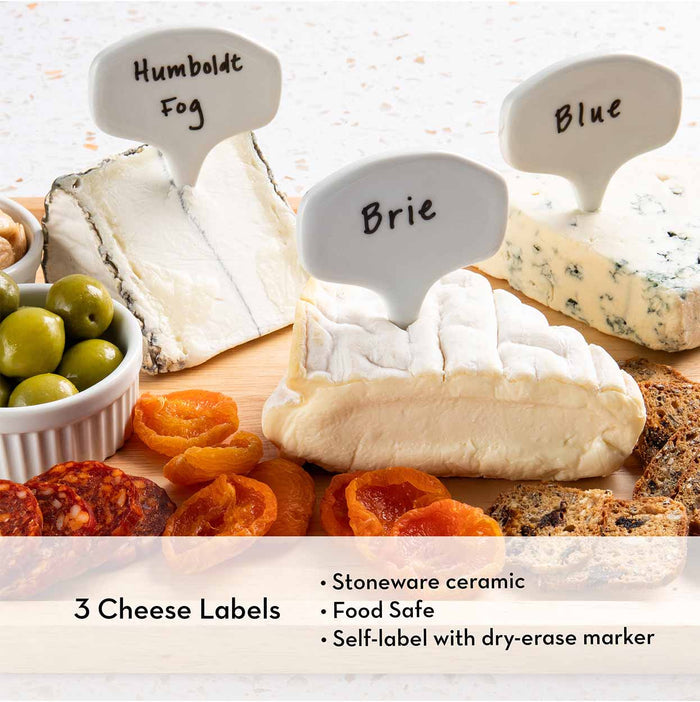 The Meg Quinn Charcuterie Board & Accessories Kit by Maple features food-safe, writable stoneware cheese labels with labeled wedges of Humboldt Fog, Brie, and Blue cheese, surrounded by dried apricots, olives, crackers, and cured meat.